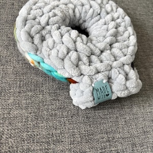 Pode incluir: Uma scrunchie cinza, feita à mão, com uma aparência espessa e texturizada. A scrunchie tem um sotaque colorido de azul, laranja e amarelo. Uma pequena etiqueta que diz "MADE WITH" está anexada à scrunchie.