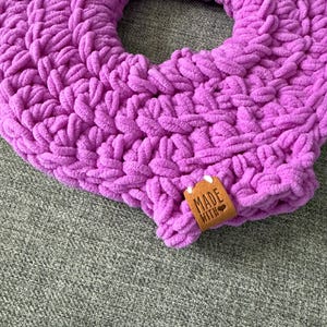 May include: A close-up of a handmade, circular, lavender-colored knitted item. The thick yarn is looped and textured, with a small brown tag that reads "MADE WITH". The item is resting on a gray, textured surface.
