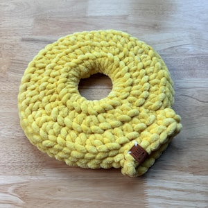 Peut inclure: Un objet crocheté en forme de donut, de couleur jaune vif. L'objet est fait d'un fil doux et texturé et possède un trou au centre. Une petite étiquette marron avec les mots "MADE WITH" est attachée.