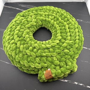 May include: A round, lime green, chunky knit cushion with a central circular opening. The soft, textured yarn is arranged in a thick, looped pattern. A small, rectangular brown leather tag is attached, featuring the text MADE WITH LOVE.