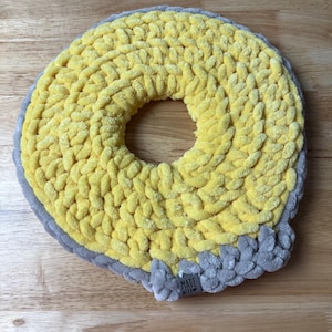 May include: A handmade, donut-shaped cushion in yellow with a gray border. The cushion is made of a thick, textured yarn and features a label that reads "MADE WITH".