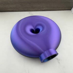 Puede incluir: Un objeto circular en forma de disco con un acabado púrpura iridiscente vibrante. La superficie presenta una textura suave y curva con una hendidura central y una abertura cilíndrica que sobresale en el borde inferior, creando una forma hueca.