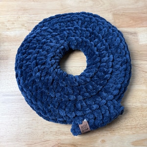 May include: A circular, dark navy blue infinity scarf crafted from thick, plush, chunky yarn. The texture is soft and voluminous with a consistent knit pattern throughout. A small, rectangular brown leather tag is attached, reading MADE WITH LOVE.