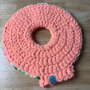 Peut inclure: Un article fait à la main en forme de donut crocheté de couleur corail avec un trou au centre. L'article a une surface texturée et un bord multicolore. Une petite étiquette bleue avec les mots "MADE WITH" est attachée.