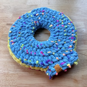 Peut inclure: Un donut crocheté bleu fait à la main avec des vermicelles colorés. Le donut a une base jaune et un trou au centre. Une petite étiquette en bois avec l'inscription "Made with love" est attachée sur le côté. Le donut est posé sur une surface en bois.