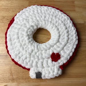 Peut inclure: Une couronne crochetée blanche faite à la main avec une bordure rouge et un petit cœur rouge. La couronne est de forme circulaire avec un trou au centre. Une petite étiquette avec l'inscription "MADE WITH" est attachée en bas.