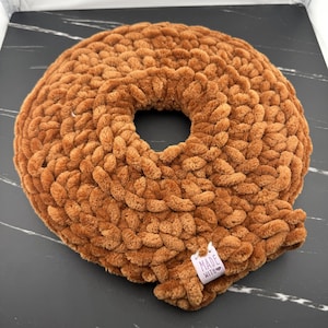 May include: A round, brown, chunky knit pillow with a soft, plush texture and a central circular opening. A small white fabric tag is attached to the bottom edge, featuring the printed text "MADE WITH" followed by a small heart symbol.