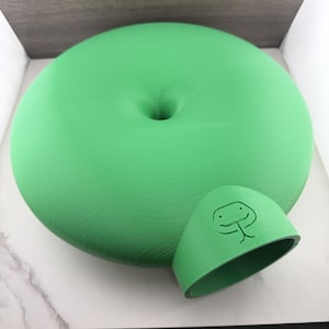 Puede incluir: Un objeto circular de color verde brillante en forma de disco con una hendidura central y un componente semicircular más pequeño adjunto que presenta un icono en relieve de una cara de serpiente estilizada con dos ojos y lengua bífida.