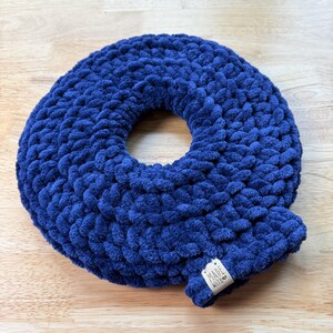 Peut inclure: Un coussin en crochet bleu marine en forme de donut. Le coussin est fait d'un fil épais et moelleux, et possède un trou au centre. Une petite étiquette avec l'inscription "MADE WITH" est attachée au coussin.