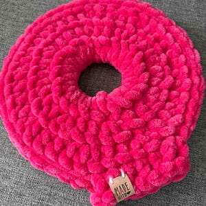 Op de afbeelding: Een felroze, handgemaakt kussen in donutvorm. Het kussen is gemaakt van dik, zacht garen, met een gat in het midden. Een klein, bruin label met de tekst "MADE WITH" is bevestigd.