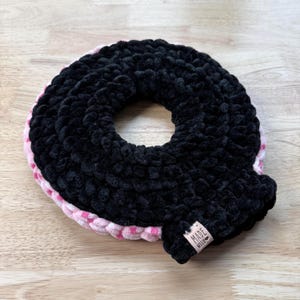 May include: A handmade, black, plush crocheted donut-shaped item with a pink and white polka-dot trim. A small, brown tag with the words "MADE WITH" is attached to the item. The item is on a light brown wooden surface.