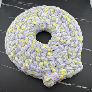 May include: A round, donut-shaped cushion made of thick, chunky yarn in a variegated pattern of light purple, soft yellow, and white. A small, rectangular fabric tag attached to the bottom edge features the text "MADE WITH" and a heart icon.