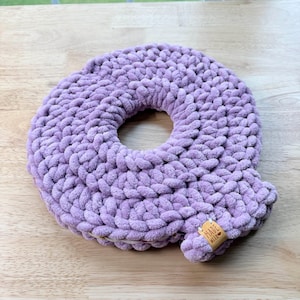 Peut inclure: Un oreiller en crochet en forme de donut de couleur lavande. L'oreiller est fait d'un fil doux et moelleux et a un trou au centre. Une petite étiquette rectangulaire avec l'inscription "MADE WITH LOVE" est attachée à l'oreiller.
