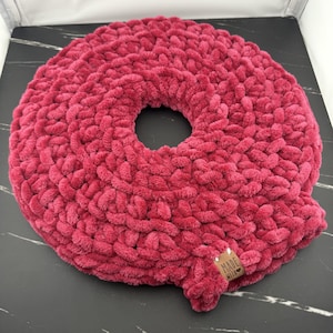 May include: A round, dark pink crochet cushion with a thick, plush, textured yarn construction and a central circular opening. A small, rectangular, light brown leather-like tag is attached to the edge, featuring the text "MADE WITH LOVE".