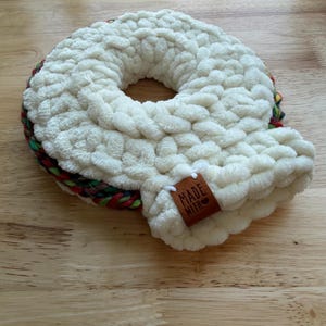 May include: A round, white, chunky knit plush ring with a soft, textured surface. The outer edge features a braided trim in variegated colors of red, green, and dark tones. A brown leather tag is attached, embossed with the text MADE WITH.