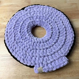 May include: A circular, chunky crochet cushion featuring a soft lavender yarn texture with a black border. The centre has a circular opening, and a small rectangular fabric tag at the bottom edge displays the text "MADE WITH LOVE".