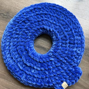 May include: A plush, royal blue donut-shaped pillow. The pillow is made of a soft, textured material and has a circular hole in the center. A small tag that says "Made With Love" is attached to the pillow.