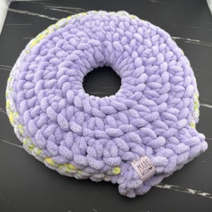 Puede incluir: Un cojín redondo con forma de rosquilla hecho de lana gruesa de color lavanda. La textura es suave y afelpada, con una abertura circular central. Una pequeña etiqueta de tela rectangular está unida al borde, impresa con el texto MADE WITH.