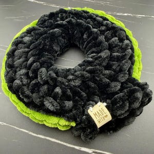 Puede incluir: Una bufanda circular de ganchillo grueso con un interior de hilo negro texturizado y un borde de hilo verde lima vibrante. Una pequeña etiqueta de tela de color claro está unida al borde, mostrando claramente el texto MADE WITH.