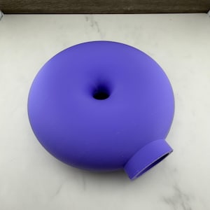 Op de afbeelding: Een glad, mat paars donutvormig object met een centrale ronde opening en een kleine, cilindrische uitstulping aan de zijkant. Het item heeft een egale, effen kleur en een afgeronde, symmetrische vorm met een platte onderkant.