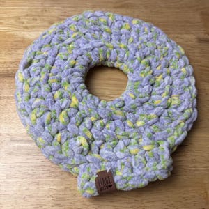 May include: A handmade, circular crochet item in shades of lavender, green, and yellow. The plush, textured piece has a central opening and a small tag that reads "MADE WITH". The item is likely a decorative or functional home accessory.