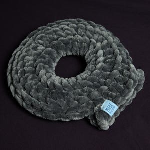 Puede incluir: Una almohada gris en forma de rosquilla, hecha de un material suave y afelpado. La almohada tiene un agujero en el centro y una pequeña etiqueta azul que dice "MADE WITH LOVE". Sobre un fondo oscuro.