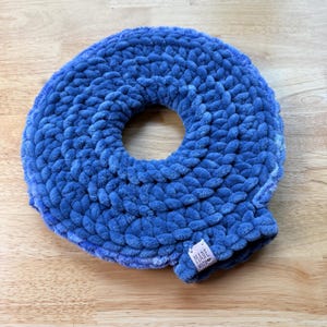 Peut inclure: Un article au crochet en forme de donut bleu fait à la main. Le fil pelucheux et texturé est d'une nuance de bleu vif. Une petite étiquette avec les mots "MADE WITH" est attachée au bord de l'article. L'article est posé sur une surface en bois.