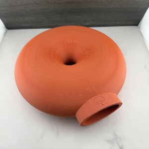 May include: A terracotta-coloured, doughnut-shaped ceramic vessel with a central circular opening and a single, protruding cylindrical spout on the side. The spout features a small, etched line drawing of a wine glass icon on its top surface.