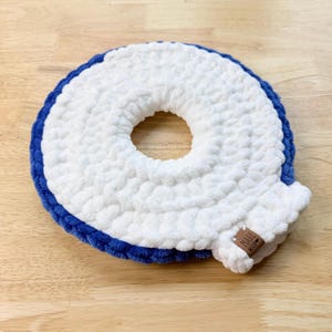 Peut inclure: Un objet fait à la main en forme de donut crocheté, blanc et bleu. Le centre est blanc, avec une bordure bleue. Une petite étiquette marron avec les mots "Made with Love" est attachée sur le côté. L'objet est posé sur une surface en bois brun clair.