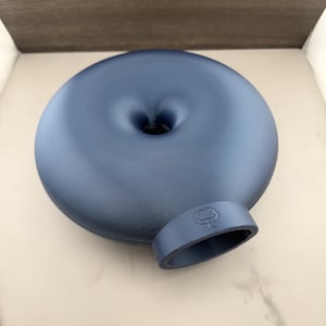 May include: A matte navy blue, donut-shaped object with a central indentation and a protruding cylindrical opening at the bottom. The opening features a small, embossed icon of a stylized creature with two eyes and a mouth, centered on the rim.