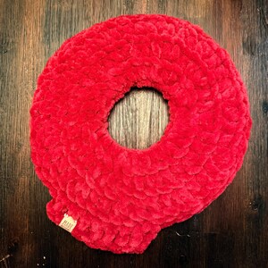 May include: A vibrant red, plush, donut-shaped pillow. The pillow is made of a soft, textured material and features a circular opening in the center. A small tag with the word "MADE" is attached to the pillow.