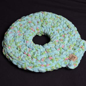 Può includere: Un oggetto circolare all'uncinetto fatto a mano, nei toni dell'azzurro, del verde e del rosa. Il pezzo in peluche strutturato ha un foro al centro e una piccola etichetta con la scritta "MADE WITH". L'oggetto è posto su uno sfondo nero.