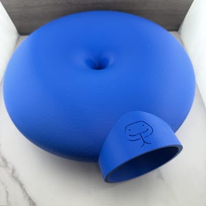 May include: A bright blue, circular, donut-shaped object with a smooth, matte finish. It features a central indentation and a protruding, tunnel-like entrance on one side, which is embossed with a simple, stylized line drawing of a snake face.