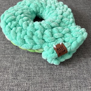 May include: A handmade, mint green scrunchie with a thick, textured knit. The scrunchie has a donut shape with a small, brown tag that reads "MADE WITH". The scrunchie is resting on a gray surface.