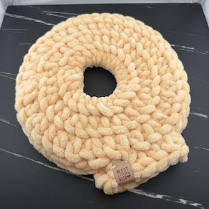 Op de afbeelding: Een rond, perzikkleurig gehaakt kussen met een dikke, grove breistructuur en een ronde opening in het midden. Aan de onderrand zit een klein, rechthoekig bruin label met de tekst "MADE WITH" en een klein hartje erop gedrukt.