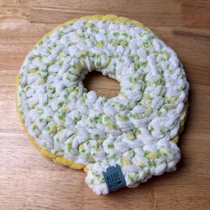 May include: A handmade, crocheted coaster in a donut shape. The coaster features a white base with green and yellow accents, and a yellow border. A small blue tag with the words "MADE WITH" is attached.