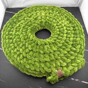Può includere: Un cuscino rotondo in maglia grossa color verde lime con un'apertura circolare centrale. La consistenza è morbida e soffice, creata con spessi anelli di filato. È presente una piccola etichetta rettangolare in pelle marrone con il testo "MADE WITH LOVE".