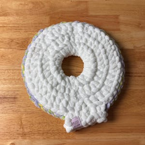 Peut inclure: Un article blanc en forme de beignet, crocheté, avec un trou central. L'article a un aspect texturé et moelleux. Les bords sont garnis de couleurs pastel. Une petite étiquette avec "MADE WITH" est attachée.