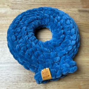 May include: A plush, blue, crocheted letter "Q" with a central hole. The letter is made of thick, textured yarn. A small, brown leather tag with the words "MADE WITH" is attached to the bottom of the letter.
