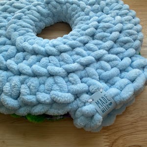 Op de afbeelding: Een rond, lichtblauw gehaakt kussen gemaakt van dik, pluche garen. Het kussen heeft een centrale ronde opening en een klein, vierkant, lichtblauw label aan de rand met de tekst "MADE WITH" en een hartje erop gedrukt.
