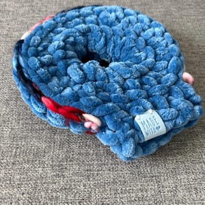 May include: A blue, plush scrunchie with a soft, textured appearance. The scrunchie has a hole in the center and a small tag that reads "MADE WITH". Accents of red, pink, and navy blue are visible.