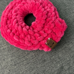 Pode incluir: Uma scrunchie de cabelo rosa brilhante, feita à mão, em forma de donut. A scrunchie é feita de um fio espesso e texturizado e apresenta uma pequena etiqueta castanha que diz "MADE WITH". A scrunchie está numa superfície cinzenta.