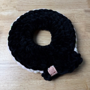 Puede incluir: Un accesorio de ganchillo circular y afelpado con una capa exterior de hilo negro grueso y una capa interior de hilo blanco con pequeños detalles verdes. Lleva una pequeña etiqueta rectangular de color marrón claro con el texto MADE WITH.