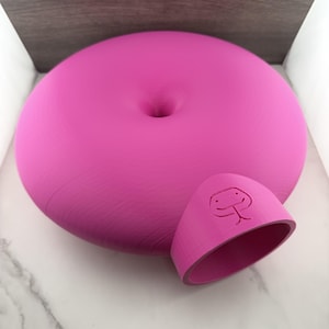 May include: A bright pink, circular, doughnut-shaped object with a central indentation. Attached to the side is a protruding, tunnel-like opening featuring an embossed, simple line-drawn face with two eyes and a mouth, resembling a stylised snake or creature.