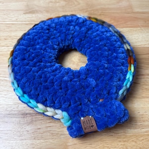 May include: A handmade, blue crocheted donut-shaped item with a multi-colored border. The item has a soft, plush texture and a small brown tag that reads "MADE WITH". The item is likely a decorative piece or a pet toy.