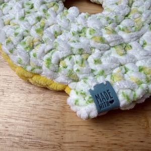 May include: A circular crochet item made of thick, plush white yarn with variegated green and yellow accents. The outer edge features a solid yellow border. A small, rectangular blue tag is attached, featuring the text MADE WITH and a heart icon.
