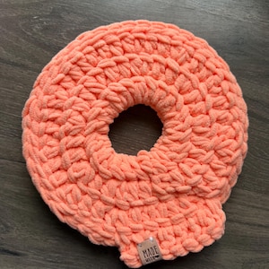May include: A coral-coloured, handmade crocheted donut-shaped item. The item is made of a soft, plush yarn and has a hole in the centre. A small tag that says "MADE WITH" is attached to the side.
