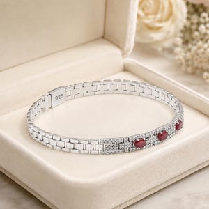 Könnte beinhalten: Ein silberfarbenes Gliederarmband mit einem 925-Stempel auf dem Verschluss. Das Armband ist mit einem Abschnitt aus kleinen klaren Steinen und drei ovalen, tiefroten Steinen verziert, die in einer Reihe auf dem Metallband sitzen.