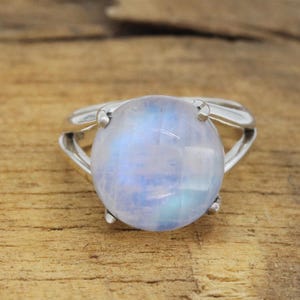 May include: A silver ring featuring a large, round, iridescent moonstone. The moonstone has a soft, milky appearance with subtle blue and white hues. The ring's band has a split design, adding a modern touch.