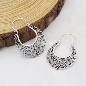 Könnte beinhalten: Ein Paar silberfarbene Creolen mit einem filigranen Design. Die Ohrringe haben eine halbmondförmige Form mit aufwendigen Wirbelmustern und einem Hakenverschluss. Die Ohrringe werden auf einer hellen Oberfläche präsentiert.